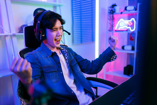 Excited young male gamer celebrates a win while playing video games on a desktop PC at night, wearing a headset in a vibrant neon-lit room, capturing gaming culture and digital lifestyle trends.