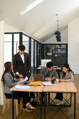 Four business professionals working together in a modern office, discussing documents and using laptops, portraying teamwork, productivity, and corporate collaboration