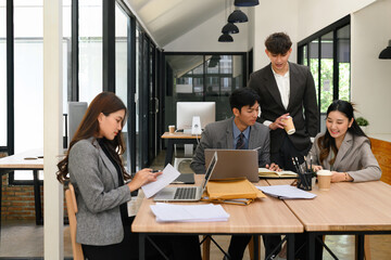 Diverse group of young professionals collaborating at a shared workspace in a modern office environment, working with laptops, papers, and coffee cups in a team meeting