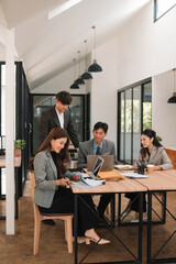 A diverse group of business people working together at a desk in a bright, modern office. Team members are engaged in discussion, reviewing documents, and using laptops