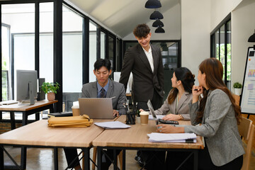 A group of professionals having a friendly conversation and working together in a modern office. One team member stands while others work with laptops and documents at the table