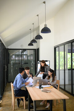 Wide-angle shot of a diverse business team in a meeting, discussing charts and working on laptops in a bright, modern office