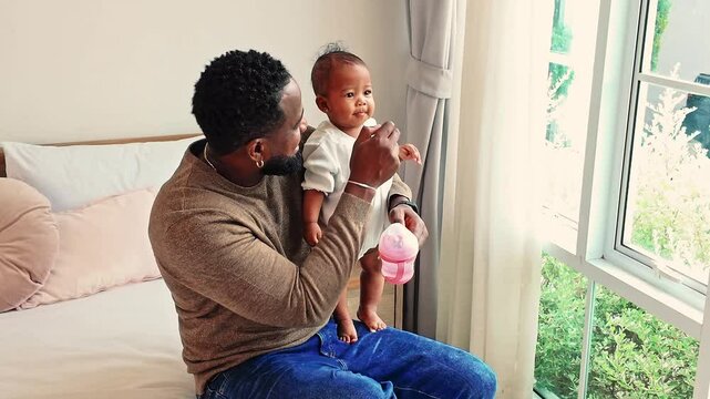 Black father holds his baby girl by a sunlit window, sharing a quiet bonding moment with a sippy cup in hand. A warm, everyday scene of modern fatherhood, parenting, and authentic family life.
