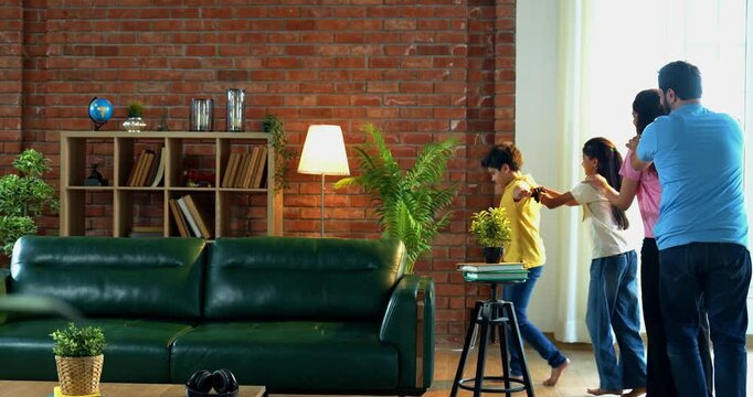 Happy Indian family playing train game indoors, little boy leading as engine while parents and sister follow behind, having fun running in a line together in cozy living room at home
