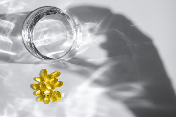 Yellow transparent capsules or pills or vitamins or supplements are near glass of water with hand shadow reaching for them on white background, representing medicine and healthcare
