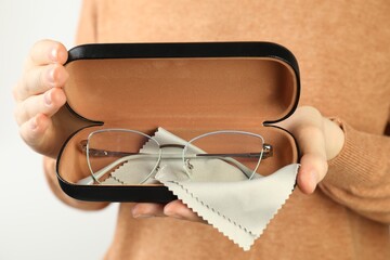 Woman holding case with stylish glasses and cleaning cloth on light background, closeup
