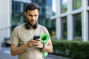 A focused athlete prepares for his fitness routine, checking his phone outdoors with a yoga mat.
