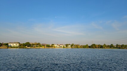 Obraz premium Serene Lakeside View Under Clear Blue Sky with Peaceful Homes and Calm Water Reflections in the Background