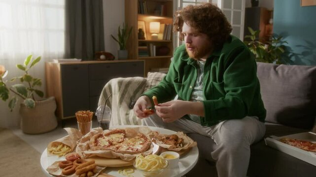 Chubby red-haired man dipping onion rings into cheese dipping sauce and ketchup, eating fast food at hame in living room