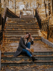 Beautiful blonde young woman in hat and coat sitting on the stairs in the autumn park.