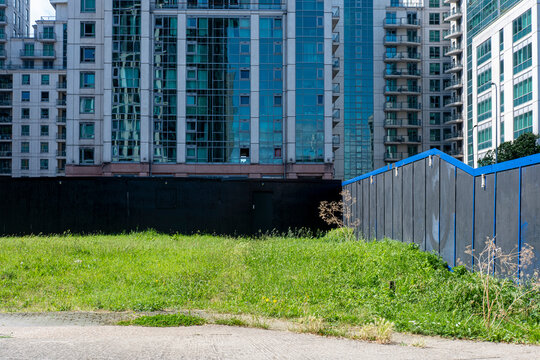 Vacant green lot surrounded by steel fence and high-rise buildings. Urban transformation, unused land, and real estate development in growing city environments captured in daylight.