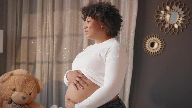 Charming pregnant african woman alone in her cozy apartment, stroking belly. Single lady waiting for baby, family and pregnancy, mother love to future baby, tenderness and care, happy motherhood