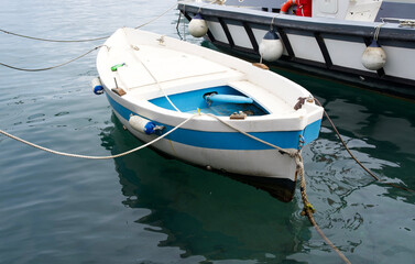 Small blue and white fishing boat anchored in calm water, reflecting the serene surroundings, with a larger vessel in the background, showcasing maritime tranquility and coastal lifestyle