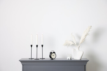 Stylish candlesticks with burning candles, other decor and alarm clock on fireplace near white wall...