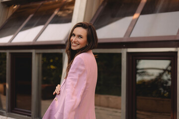 Happy woman walking outdoor on the street turn around, smiling. Brunette long hair girl wear long sleeve top and stylish pink blazer. Positive emotions, fashion outfit.