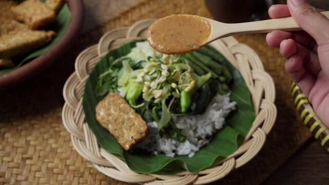 A hand drizzling peanut sauce on pecel ingredients