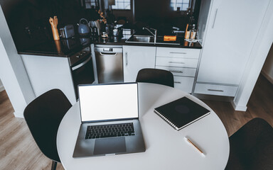 A modern kitchen setup with a round white table, open laptop with screen mockup, notebook, and pen. The background features sleek kitchen appliances and cabinets, creating a clean minimalist workspace