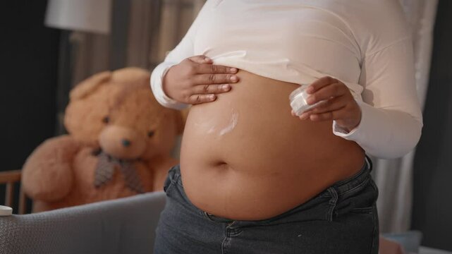 Safe and hypoallergenic cosmetics for pregnant women, closeup view of abdomen. Natural organic extracts in cream for prevention of stretch mark and moisturizing skin, african woman massaging her belly
