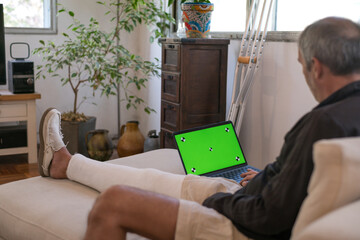 Man with a broken leg working at home on a green screen laptop