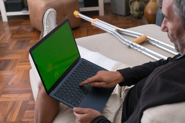 Man with broken leg using laptop with green screen at home