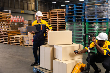 Warehouse team collaborates as one worker crouches to scan boxes with handheld scanner while colleague updates data on laptop, ensuring cargo tracking in industrial aisle. Diverse males, mid-30s