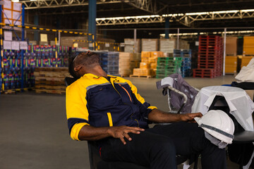 Fototapeta premium Exhausted warehouse worker exhausted in chair, tired eyes closed holding safety helmet working late, overload, shift break in logistics center, African male in yellow vest, industrial racks industry