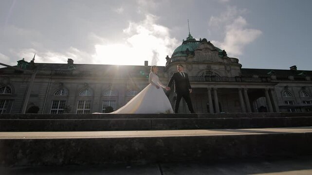 Bride and groom holding hands on stone steps in front of historic building, elegant wedding scene in sunlight