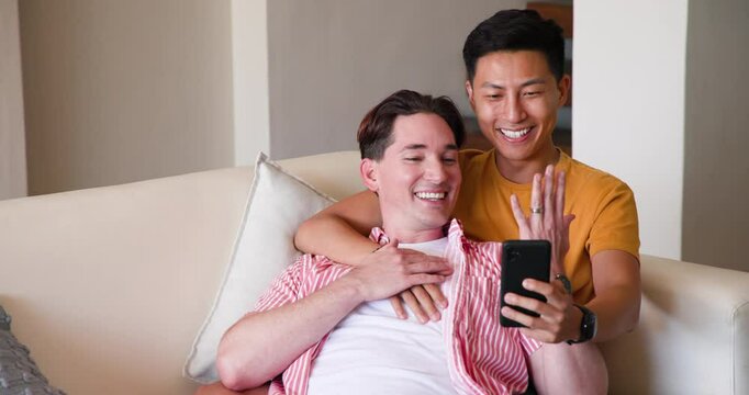 Diverse gay couple relaxing on couch, smiling and looking at smartphone together at home, copy space
