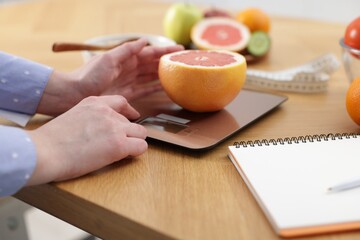 Woman weighting grapefruit on kitchen scale at wooden table, closeup