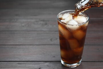 Pouring refreshing cola from bottle into glass at wooden table, closeup. Space for text