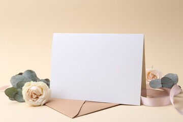 Blank greeting card, envelope, eucalyptus leaves, roses and ribbon on beige background, closeup