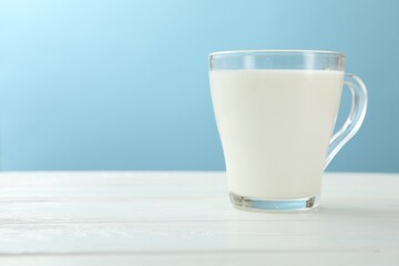 Fresh milk in glass cup on white wooden table against light blue background, closeup
