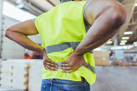 Worker Experiencing Back Pain in a Warehouse Environment Wearing High-Visibility Vest - Powered by Adobe