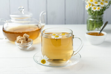 Delicious chamomile tea in glass cup, brown sugar, teapot, jam and fresh flowers on white wooden table, closeup