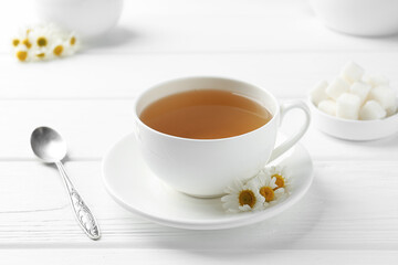 Delicious chamomile tea in cup, fresh flowers, sugar cubes and spoon on white wooden table, closeup