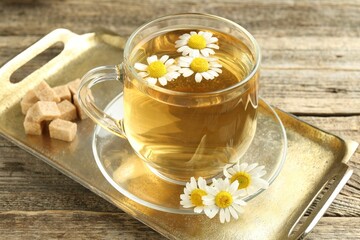 Delicious chamomile tea in glass cup, fresh flowers and brown sugar on wooden table, closeup