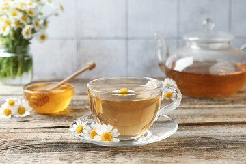 Delicious chamomile tea in glass cup, fresh flowers, honey and teapot on wooden table, closeup