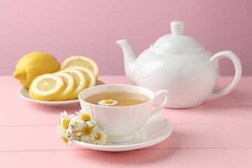 Delicious chamomile tea in cup, fresh flowers and lemon on pink wooden table, closeup
