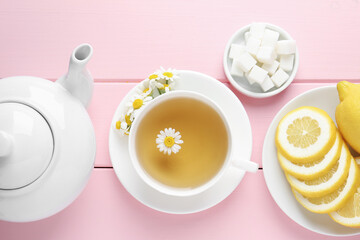 Delicious chamomile tea in cup, fresh flowers, sugar cubes and lemon on pink wooden table, flat lay