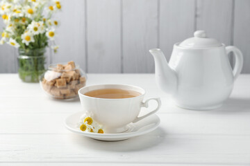 Delicious chamomile tea in cup, fresh flowers and brown sugar on white wooden table