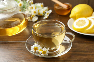 Delicious chamomile tea in glass cup, fresh flowers, lemon and honey on wooden table, closeup