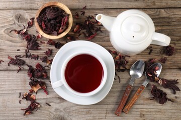 Delicious hibiscus tea in cup, teapot, dry roselle sepals and spoons on wooden table, flat lay