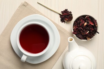 Delicious hibiscus tea in cup, teapot, dry roselle sepals and spoon on light wooden table, flat lay