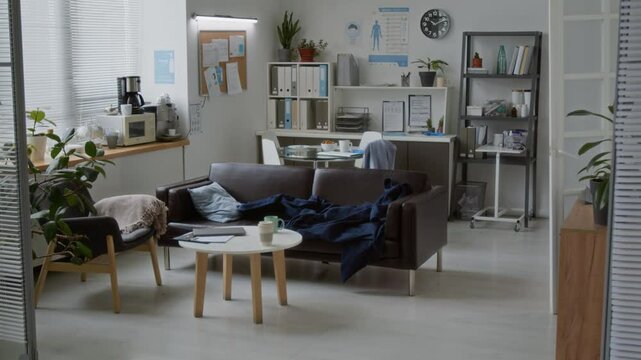 Part of empty medical office with leather sofa, armchair and small round table against workplace of clinician standing by shelves with folders and medical documents