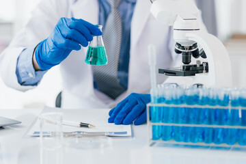 Fototapeta premium Scientist analyzing liquid in laboratory setting. Scientist is holding a conical flask with a blue liquid, conducting research in a laboratory equipped with a microscope and test tubes.