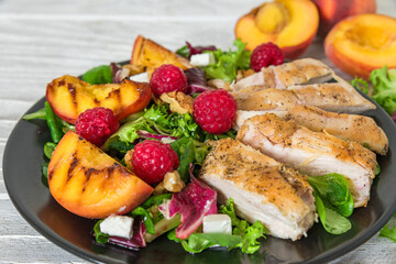 Grilled chicken breast salad with peaches, feta cheese, raspberries, walnuts and mixed greens on black plate on white wooden background. Close up. Healthy diet food. Summer dinner