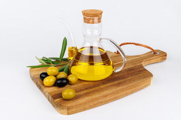 Glass oil cruet with olive oil, green olives, and olive branches on a dark cutting board over white background