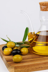 Glass olive oil cruet with green olives and olive branches on a dark wooden cutting board, white background
