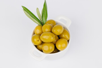 Fresh green olives in white ceramic bowl with olive branch on white backdrop