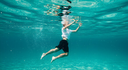 Underwater Shot of a Person Swimming in Tranquil Turquoise Water with Refracted Sunlight Wearing a White Shirt and Black Shorts